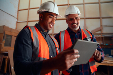 Men in reflective vest and safety helmet smiling while looking at digital tablet in woodworking factory