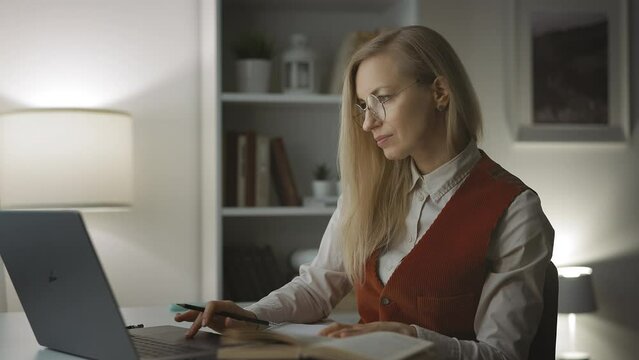 Attentive middle-aged lady learning foreign language, using laptop and paper book, maing text translation