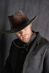 Cropped close-up shot of a man in a wide-brimmed leather cowboy hat pulled down with an adjustable cord. A man in black clothes and in a western hat is standing on a dark background. Front view.