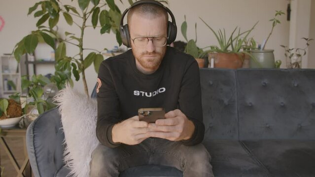 Medium close front angle of a man sitting on a couch scrolling and swiping and typing on an Iphone.