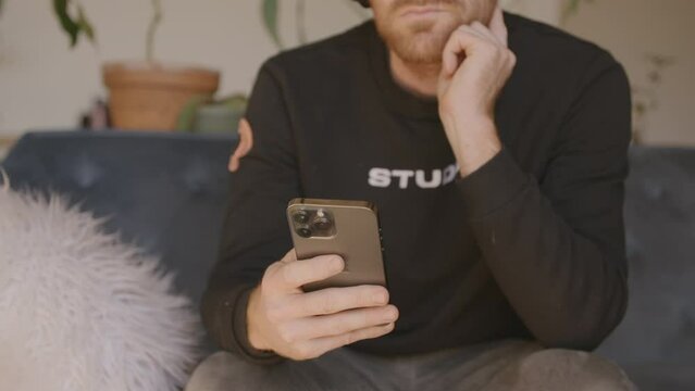 Medium close front angle of a man sitting on a couch scrolling and swiping and typing on an Iphone.