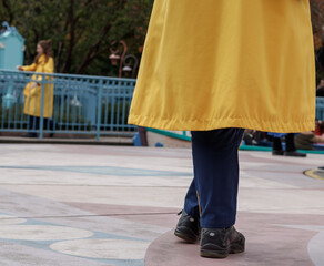 Close up on the crossed legs of a woman in yellow anorak and blue pants. Another woman is visible, blurred in the background.