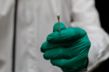 Close up on the gloved hand of a nurse holding a covid test swab. The white coat of the doctor is...