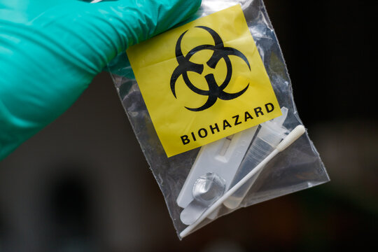 Close up of a bag marked "Biohazard" containing a used covid self test. The bag is held by a gloved doctor's hand. The dark background is blurred, focus on the bag.