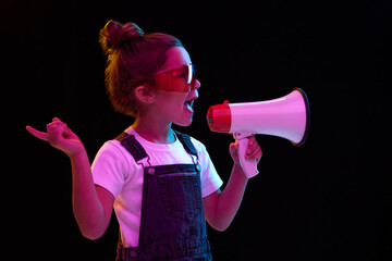 Stylish little girl, pupil in white t-shirt and denim dress shouring at megaphone isolated over dark background in neon light. Concept of beauty, kids fashion, children emotions