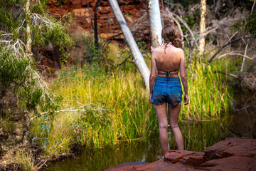 A beautiful girl in a bikini stands on red rocks above a river in a canyon in karijini park,...