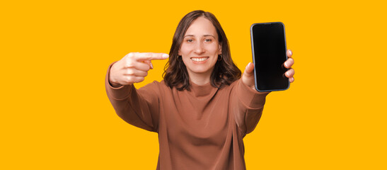 Image of young smiling woman pointing at smartphone over yellow background