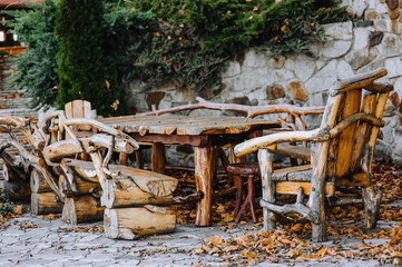 Large homemade vintage wooden products: a table, a chair, a bench stand in the park in autumn in nature with yellow maple leaves against the background of a stone wall. Photography of nature.