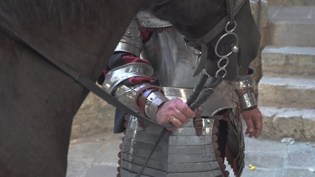 A knight in metal armor holds the reins of a horse and hands them to a woman in front of the castle walls