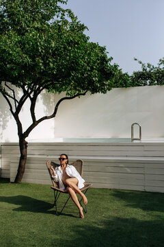 Tanned girl in a white shirt on a naked body. A woman in a white shirt, wearing sunglasses sits on a green grass or lawn. Beach summer vacation, holiday season. Backyard of the house