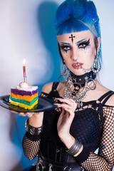 Punk girl with blue clouds, piercing and bright makeup holds a piece of colorful cake in her hands