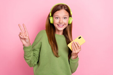 Photo of pretty cute schoolgirl wear green sweatshirt earphones chatting modern gadget showing v-sign isolated pink color background