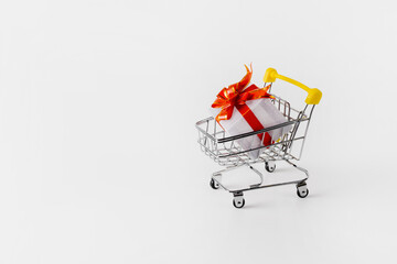 Metal cart from a supermarket with a gift on a white background. The gift is wrapped in white paper, tied with a red ribbon. The concept of buying gifts before the holiday, Christmas