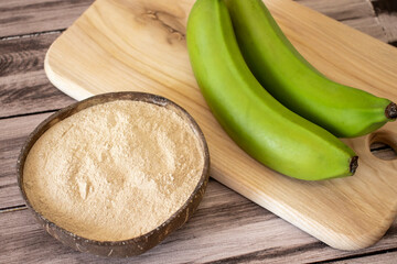 Banana brown flour in a natural bowl on a wooden surface with fresh green bananas