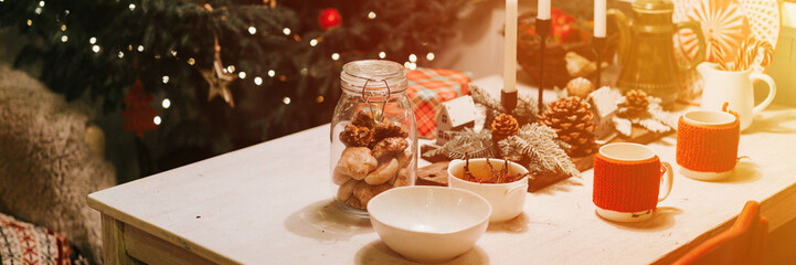 tablescapes and christmas and new year time. decorated festive cozy table with mugs, gingerbread dinner food in a jar and candles near the christmas tree. winter holiday season at home. banner. flare
