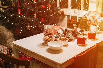 tablescapes and christmas and new year time. decorated festive kitchen cozy table with mugs, gingerbread dinner food in a jar and candles near the christmas tree. winter holiday season at home. flare