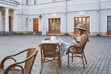 beautiful stylish young woman in a hat sits at a table in a cafe on the street, and looks at something in a smartphone