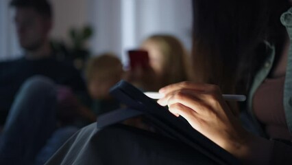 Close-up of a young hispanic woman graphic designer working on a tablet while sitting on the couch with her children. Graphic designer draws with a stylus on a tablet at night in the dark