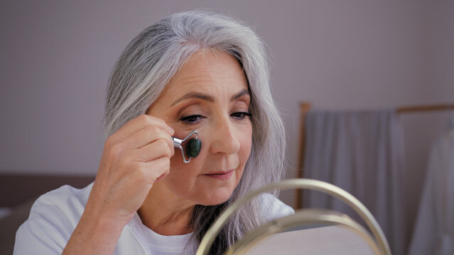 Close-up Old Gray-haired Caucasian Woman Model Grandma Elderly Lady Looking In Mirror Smooth Out Wrinkles On Face With Quartz Stone Roller Massager Rubs Lymph On Skin Doing Facial Rejuvenation Massage