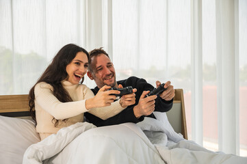 Caucasian couple holding game controller sitting on their bed playing game together having fun.	