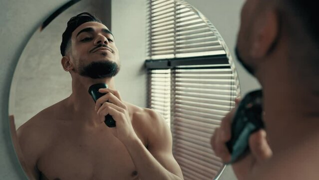 Closeup male reflection in mirror in bathroom unshaven with bristles on face shaving at home. Indian Latina handsome 30s guy bearded Hispanic man uses electric razor electrical trimmer shaver to shave