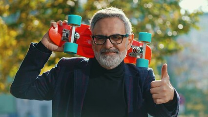 Grey-haired middle-aged white man wearing glasses and blazer standing at park holding red skateboard on his shoulder looking into camera smiling and showing thumbs up. Fun grandpa. Horizontal video - Powered by Adobe