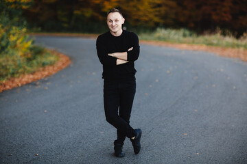 Fototapeta premium Young attractive man is standing on road in autumn forest. Guy with short hair wearing in black is looking in camera in park with yellow leaves trees. Cozy day on countryside