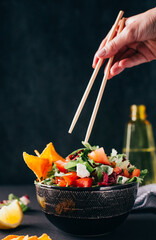 closeup of a vegetable salad in a plate with a female hand holding Chinese chopsticks on a dark background