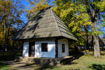 Fototapeta na wymiar Traditional Romanian house surounded with many old trees with green, yellow, orange and brown leaves in Village Museum in Herastrau Park in Bucharest, Romania in a sunny autumn day