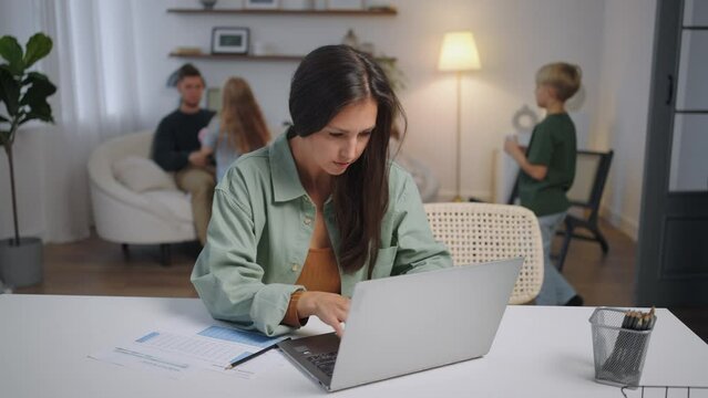A business woman works remotely at home on a laptop while her family is singing in the background . Busy business woman. Father and children play