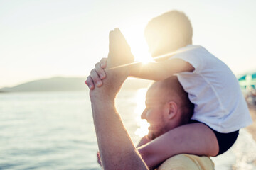 Young family having fun outdoors at the beach. Son on sitting on fathers shoulders along beach. A...