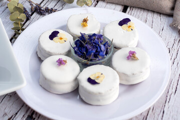 Handmade black and white chocolate alfajores with dulce de leche on plates decorated with dry edible flowers of many colors on a wooden table