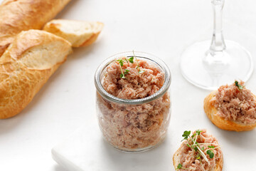 Healthy tuna pate or rillettes in a glass jar on marble board and white background. Toast with tuna on baguette.