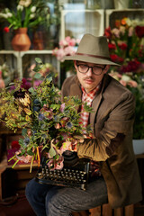 Tired or thinking emotion. Attractive caucasian male person in eyeglasses and hat sitting with old typewriter in flower shop. Poet or writer working concept. High quality vertical image