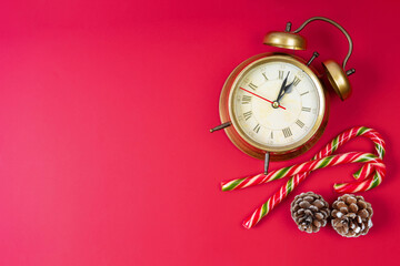 Bronze alarm clock, candy cane and fir cones on red background. Clock hands show twelve, midnight. Top view, flat lay with copy space, banner, header. Chrismas and New Year background