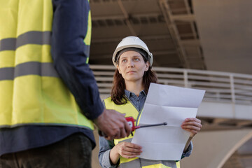 two engineers, a man and a woman in helmets and protective vests, talk on the radio with workers....