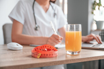 Close up view. Apple juice and measuring length is on the table. Young beautiful female doctor with laptop