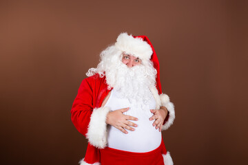 Funny fat Santa Claus keeps his hands on his stomach. Christmas eve celebration. Brown background.