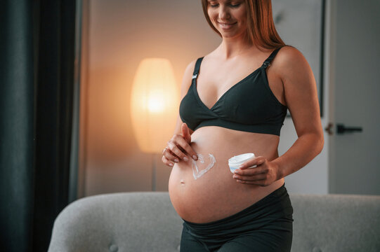 Making heart shaped picture by cream on the belly. Beautiful pregnant woman is indoors at home