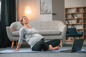 On yoga mat with laptop. Beautiful pregnant woman is indoors at home