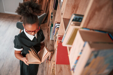 Standing and reading the book. Cute african american girl in school uniform is at home library