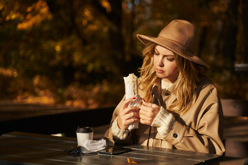 a beautiful young woman sits in an autumn park on a sunny day at a table and eats shawarma or kebab. street food, park food, street food