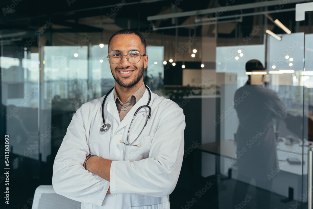 Wall mural portrait of a young handsome male hispanic doctor. standing in the glass hall of the hospital wearin