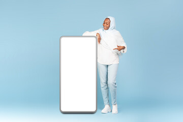 Wow, great offer. Full body length of happy black muslim woman pointing at big blank smartphone display, mockup