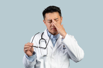 Tired sad middle aged chinese man doctor in white coat takes off glasses, rubs eyes with hand