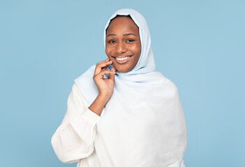 Positive african american woman in hijab looking at camera and smiling, posing on blue background and touching her face