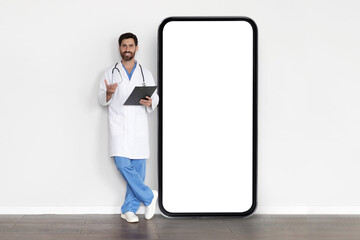 Telehealth Concept. Handsome Male Doctor In Uniform Standing Near Big Blank Smartphone