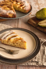 Still life on a wooden kitchen table: pear pie and a piece of homemade pear pie on a ceramic plate. Open pear pie. Vertical photo. Selective focus. Photo in rustic style.