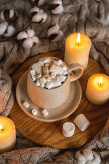 Obraz na płótnie Canvas Cozy home still life: hot chocolate or cocoa with marshmallows on a wooden board, a wool sweater, candles and a sprig of cotton. The concept of wishing a cozy evening. Selective focus.