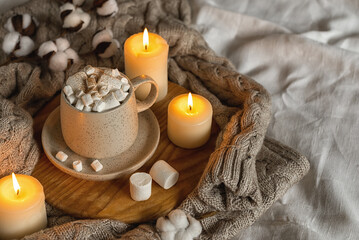 Fototapeta na wymiar Cozy home still life: hot chocolate or cocoa with marshmallows on a wooden board, a wool sweater, candles and a sprig of cotton. Horizontal photo.The concept of wishing a cozy evening. Selective focus